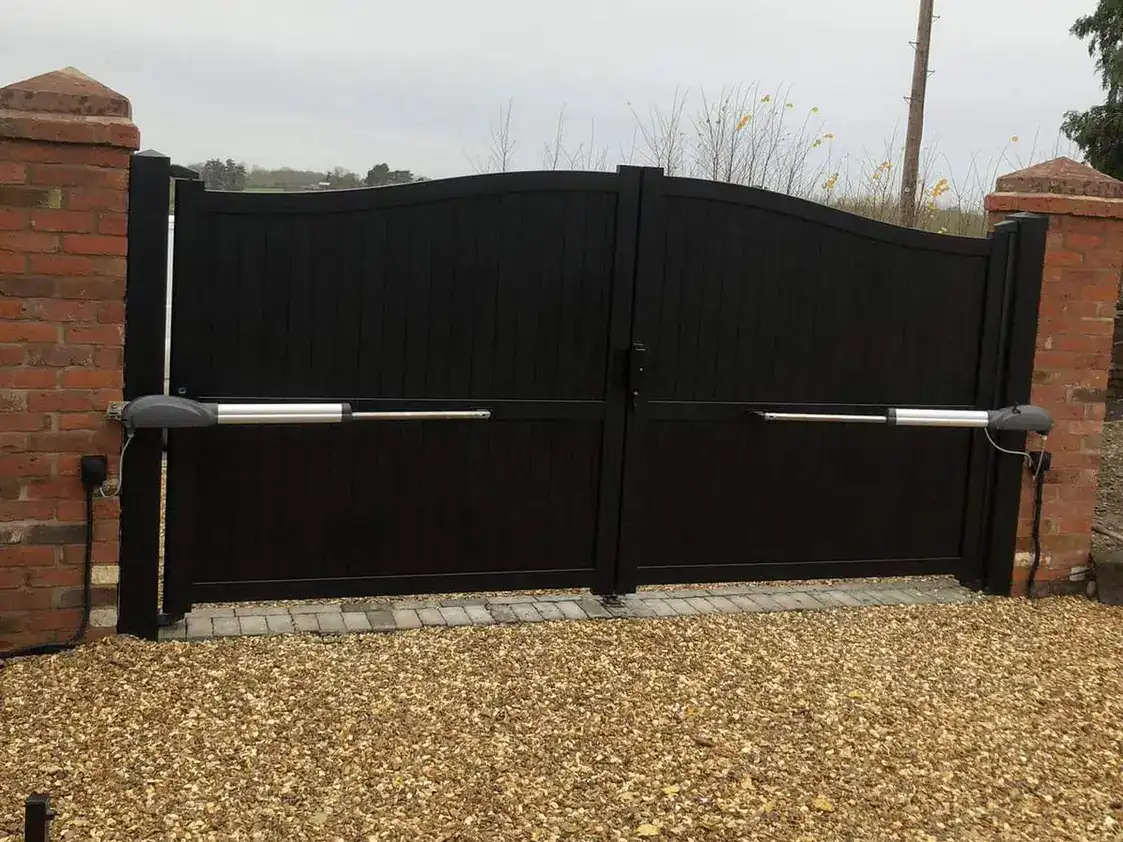 Black solid-panel driveway gates with ram arms between brick piers on a gravel drive