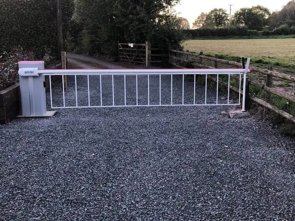 White barrier arm across a gravel track between hedges, a field gate beyond