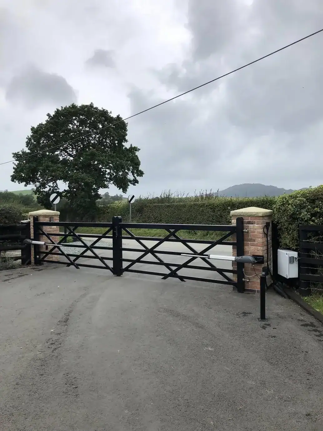 Black five-bar gates with ram arms between brick piers, an oak tree and hills beyond