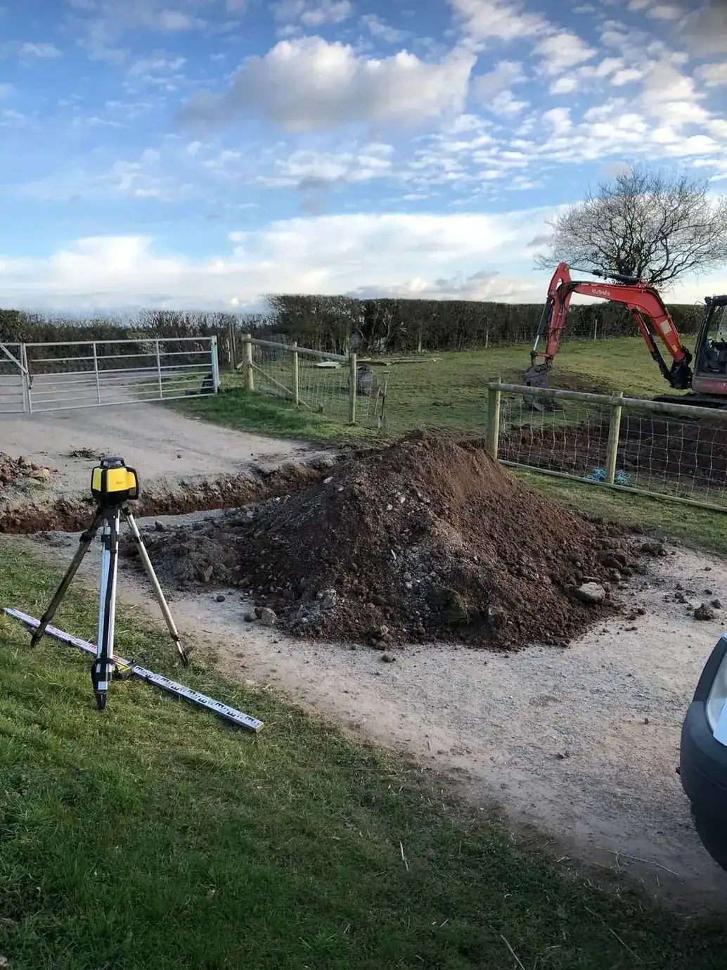 Laser level on a tripod beside a spoil heap and mini digger at a farm entrance during groundwork