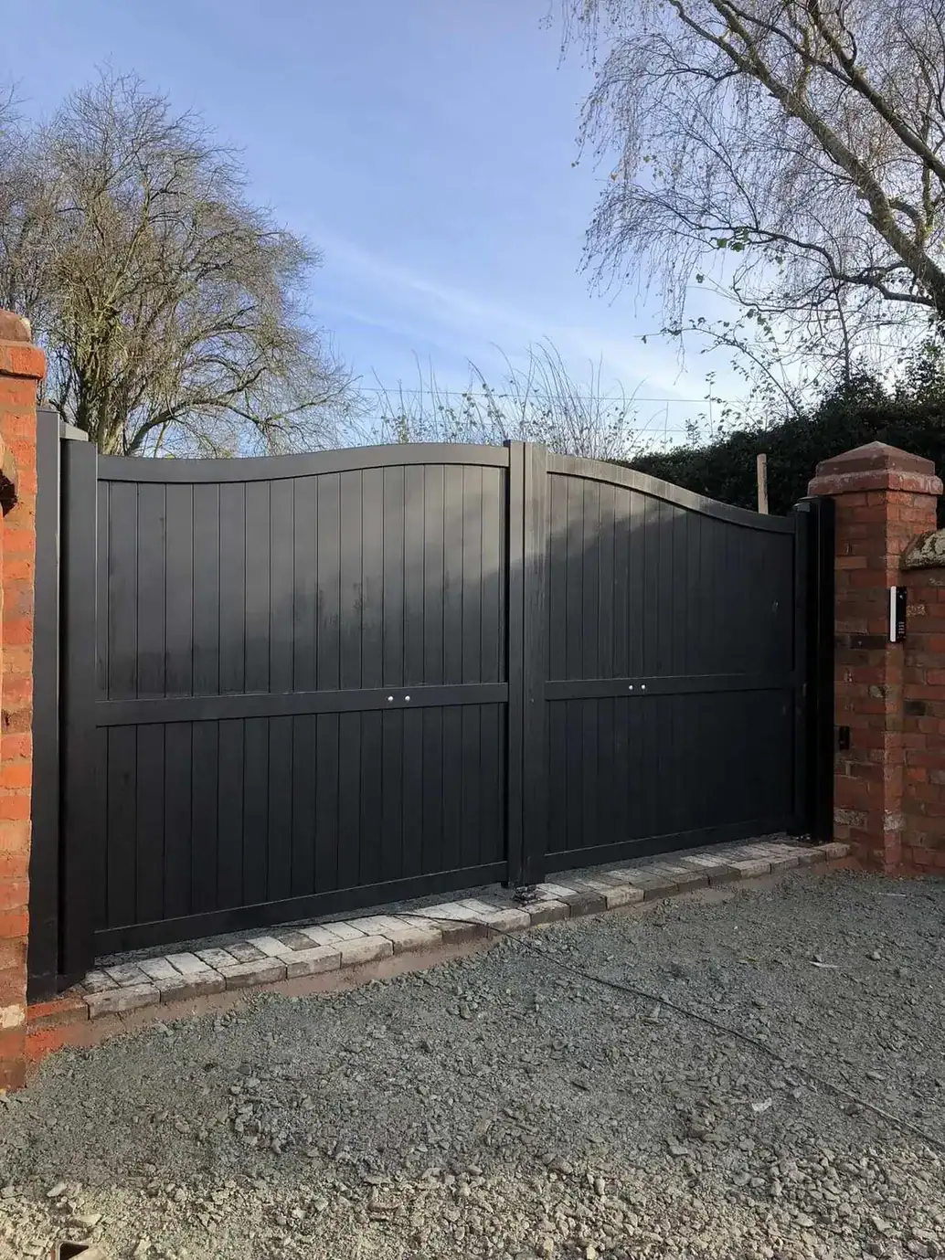 Black arched solid-panel driveway gates with a ram arm, between brick piers