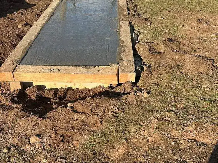 Freshly poured concrete strip in timber shuttering beside a field fence