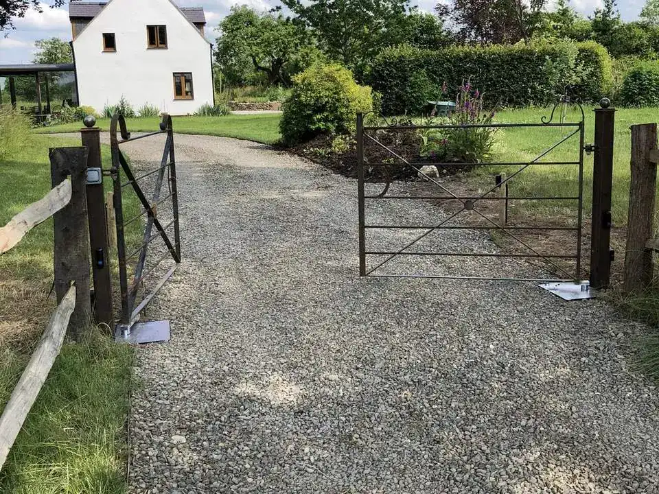 Metal field gate on timber posts across a gravel track, a white cottage behind