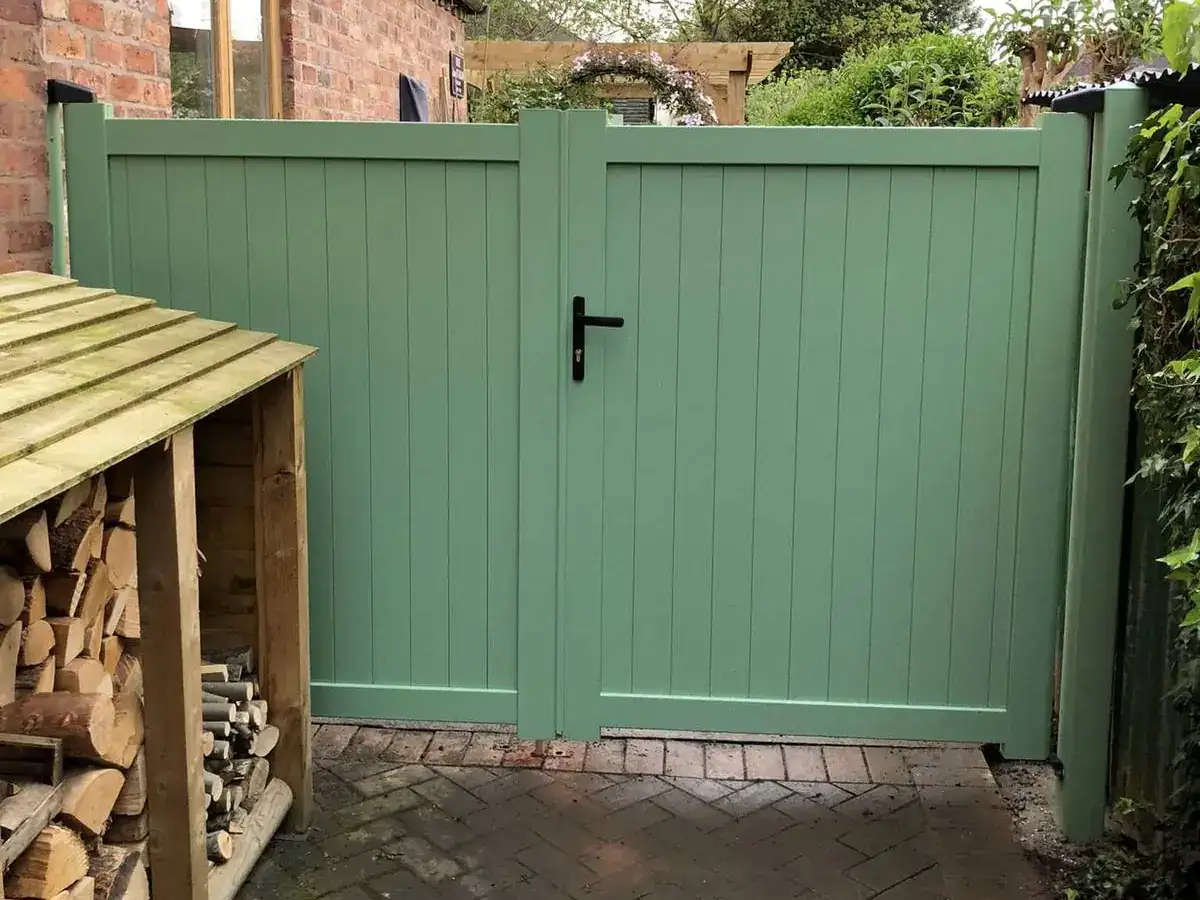 Sage green garden gates beside a log store, block paving in front