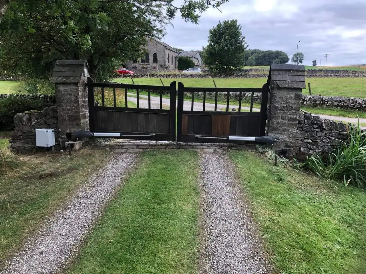 Dark timber driveway gates with ram arms, between stone piers at the end of a grass-centred track
