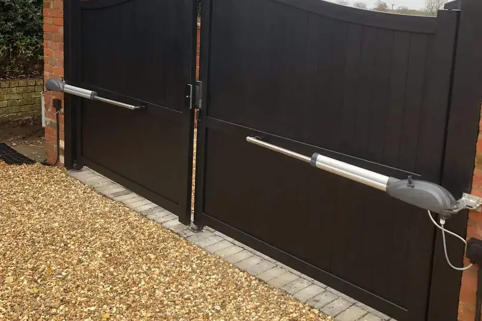 Black solid-panel swing gates with ram arms, between brick piers on a gravel drive