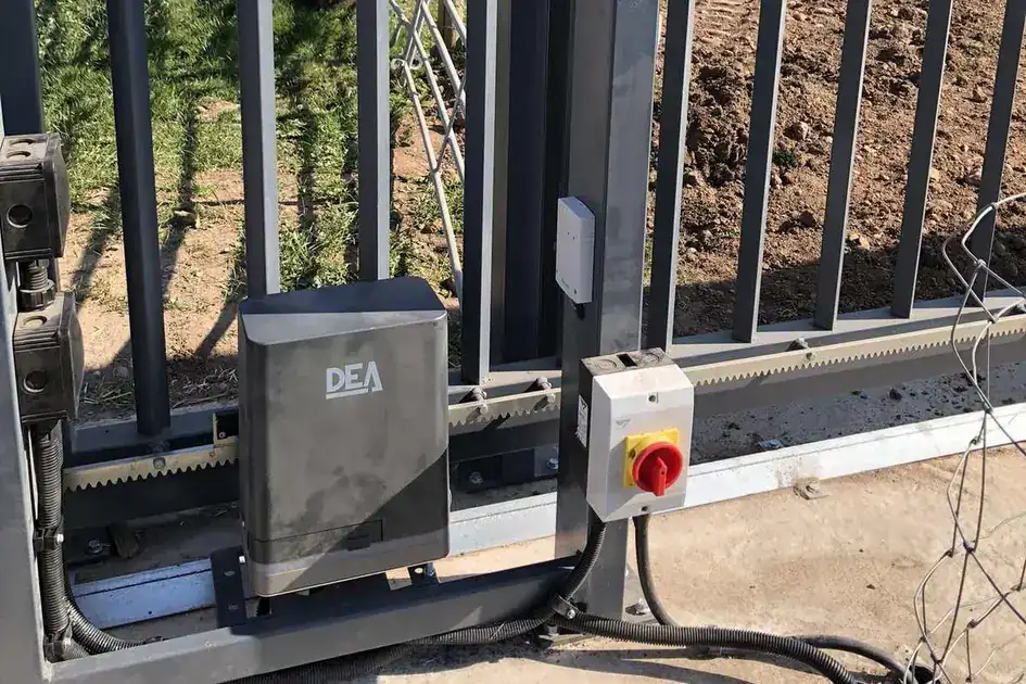 Sliding gate motor and toothed rack at the foot of a black steel sliding gate, isolator switch beside it