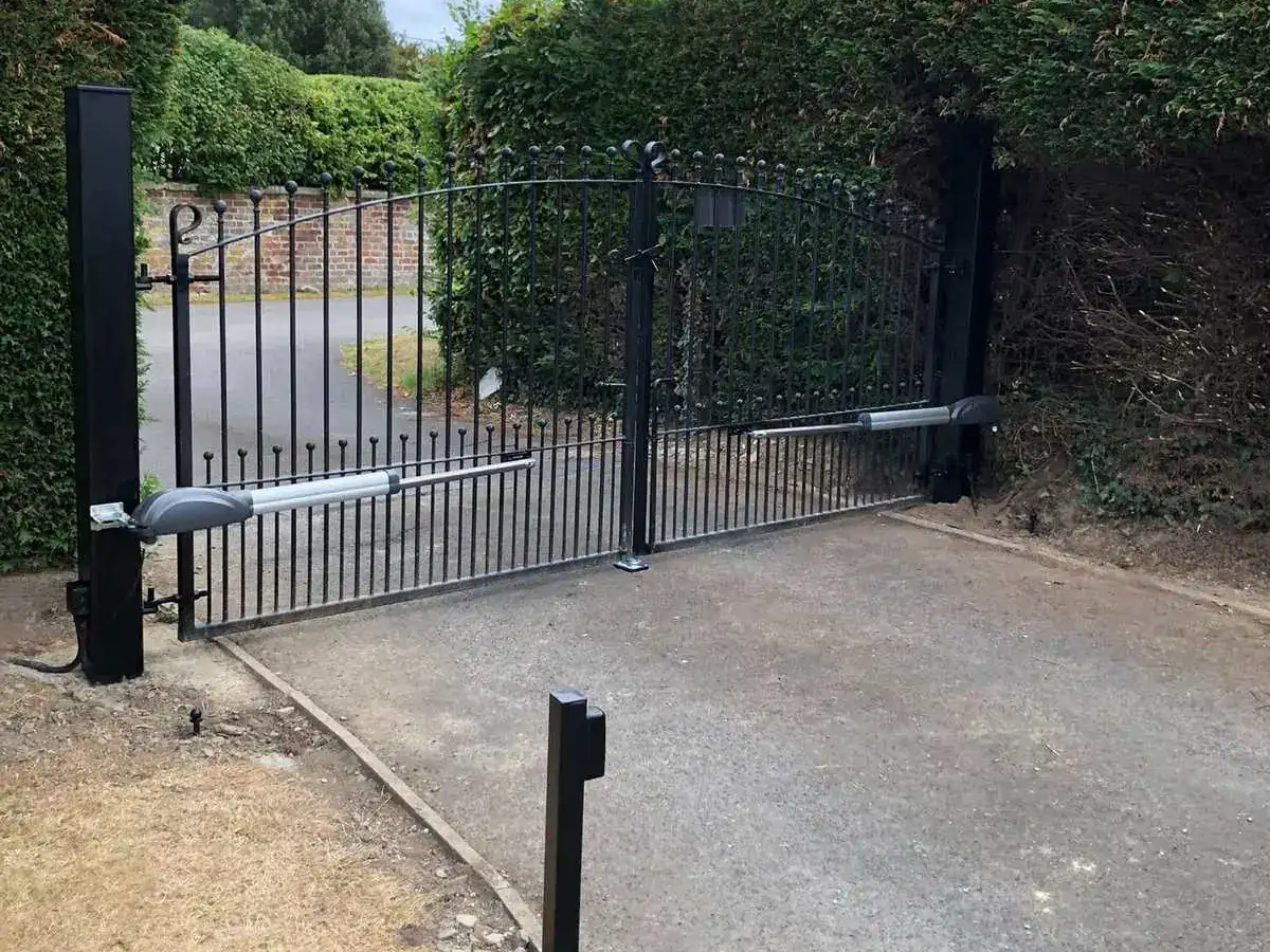 Automated iron gates between hedges, with ram arms and a photocell post either side of the drive