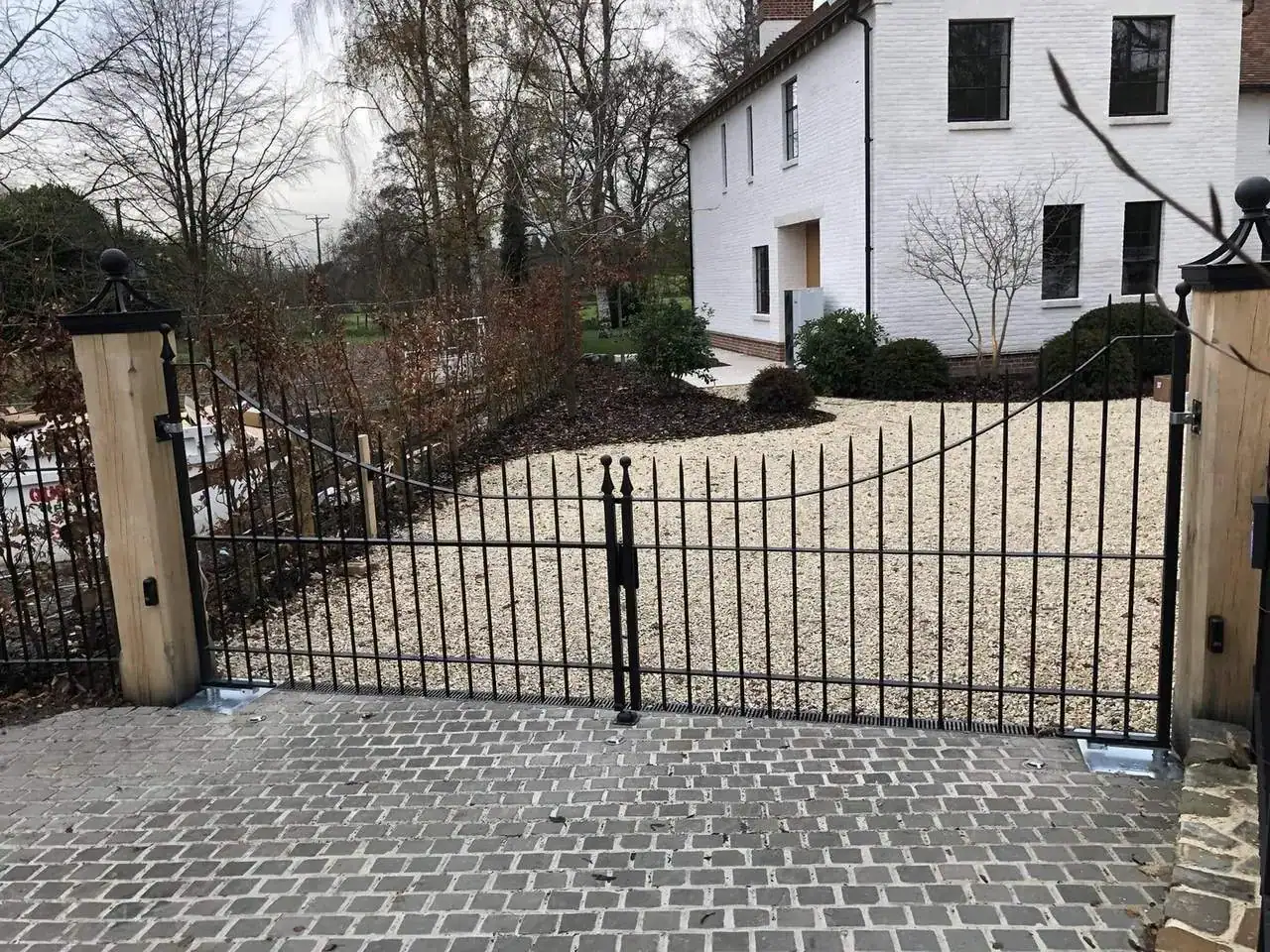 Black iron driveway gates on oak posts across a cobbled drive, white house behind