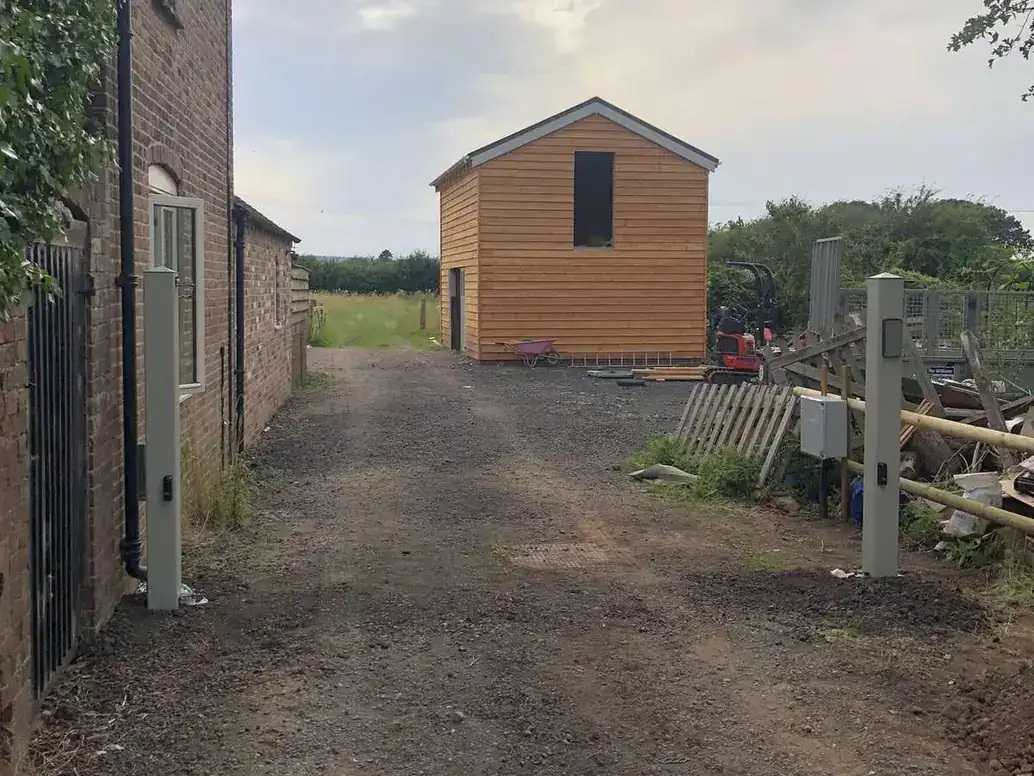 Gravel lane between a brick house and a timber outbuilding, a new gate post on the right