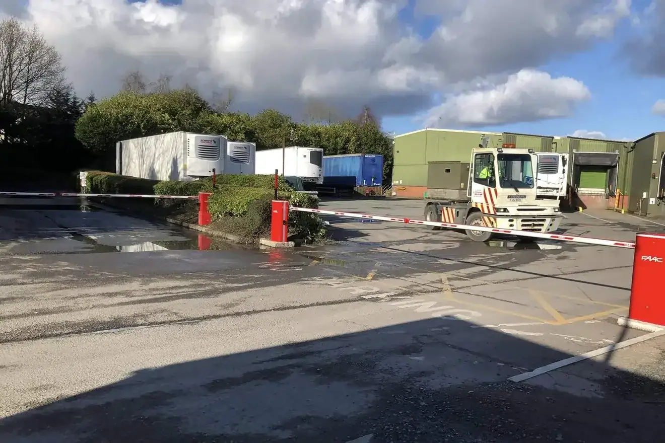 Red automatic barrier at a factory yard entrance, lorries and units behind