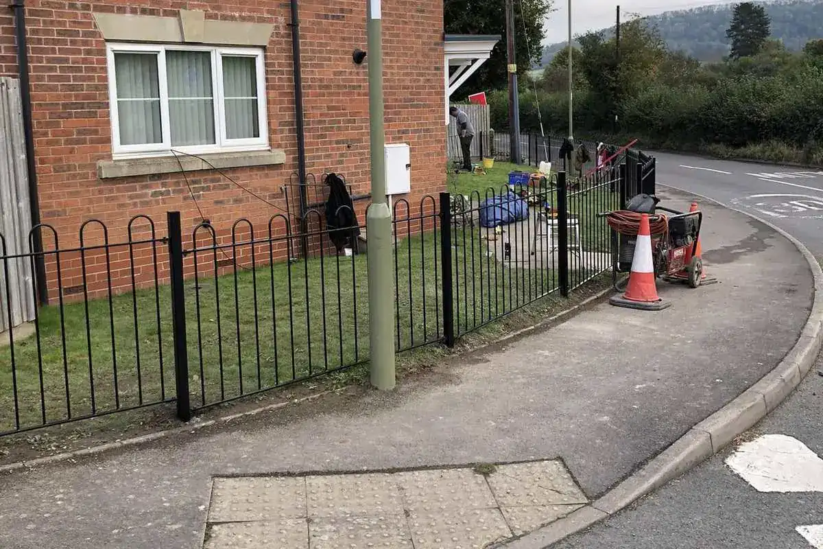 Black bow-top railings along a front lawn beside a pavement, brick house behind