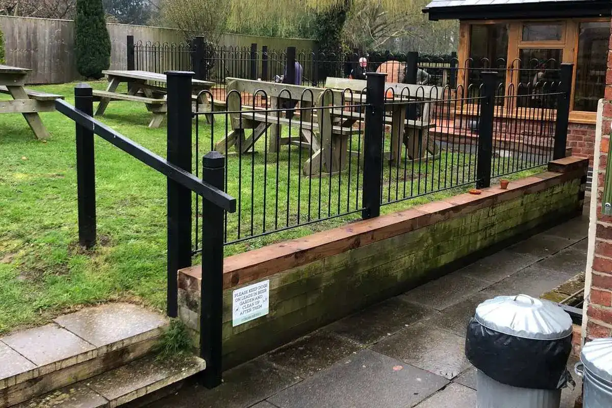 Black railings around a pub garden with picnic benches, a willow tree behind
