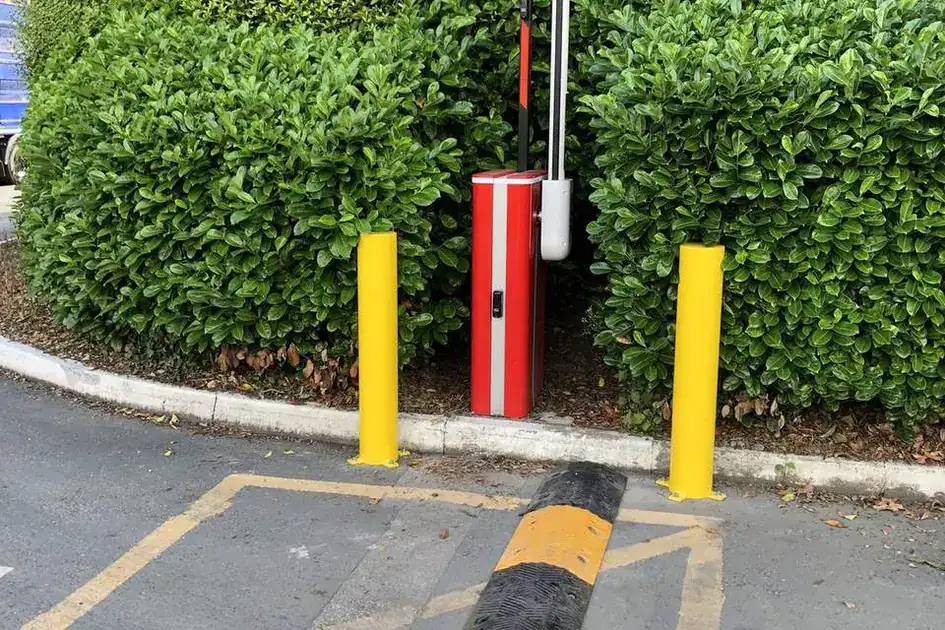Red barrier with its arm raised between two yellow bollards, a speed bump in front