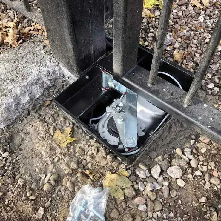 Underground gate operator in its open foundation box at the foot of an iron gate post