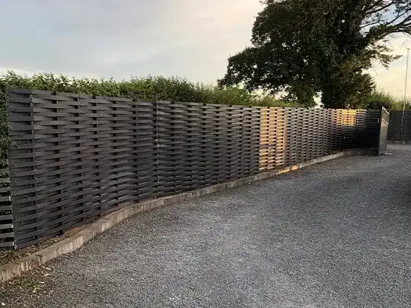 Long run of dark grey woven steel fence following the curve of a gravel drive at dusk