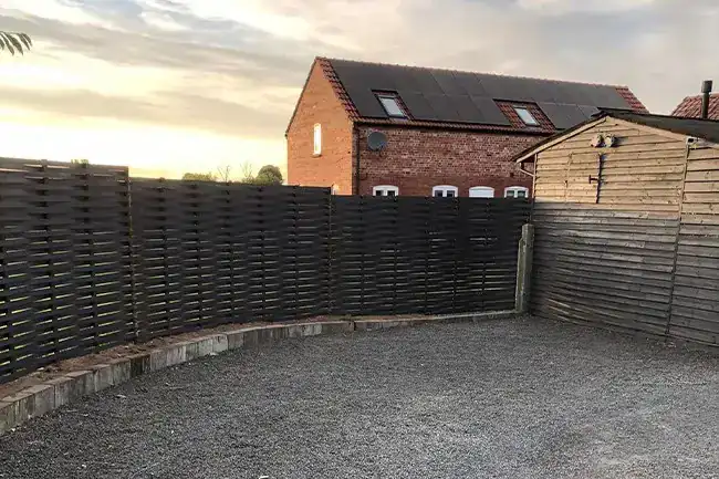 Woven steel fence curving around a gravel parking area at dusk, a brick house behind