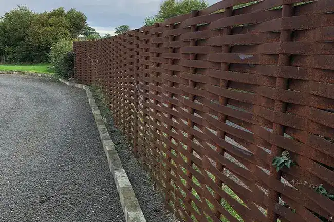 Rust brown woven steel fence running beside a gravel drive