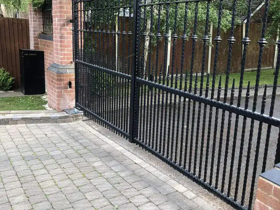 Black iron gate with an arched top and finials beside a brick pier on a block-paved drive